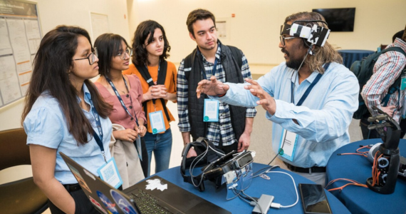 a group of students gathered around a presenter with a head piece on gesturing